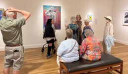 A group of older adults gather around a museum docent who is describing a painting.