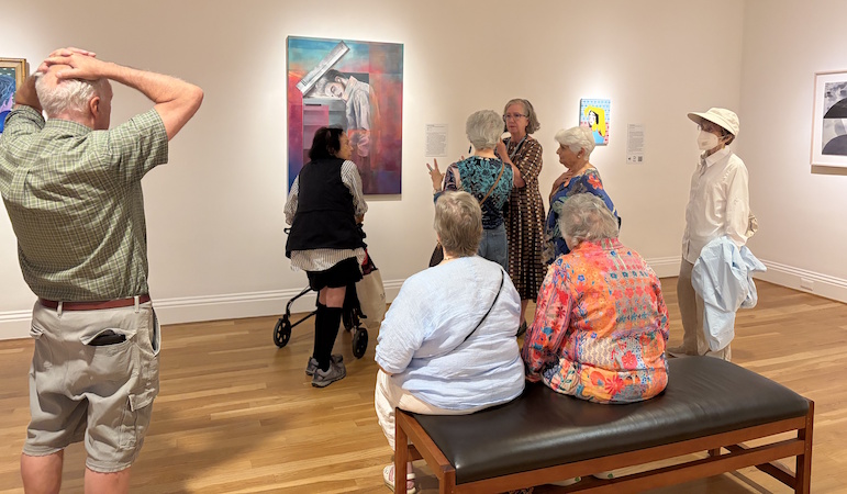 A group of older adults gather around a museum docent who is describing a painting.